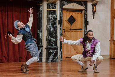 Dromio of Syracuse on tiptoes with hands upraised and leaning back crying out, wearing blue vest and hat, blue striped kneepants with white stockings and work boots as Antopholus of Syracuse squats down, looking at the audience while pointing at Dromio and wearing a fancy purple vest over white shirt and cream kneepants with white stockings and stylish shoes.